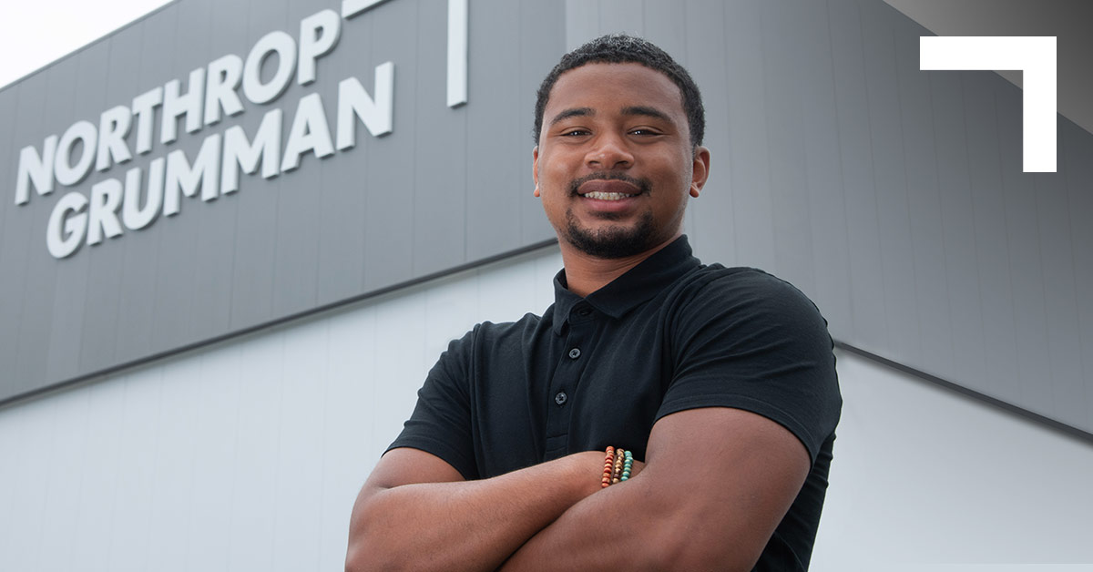 a Black male standing in a black shirt in standing in front of an office building with a Northrop Grumman sign in white letters