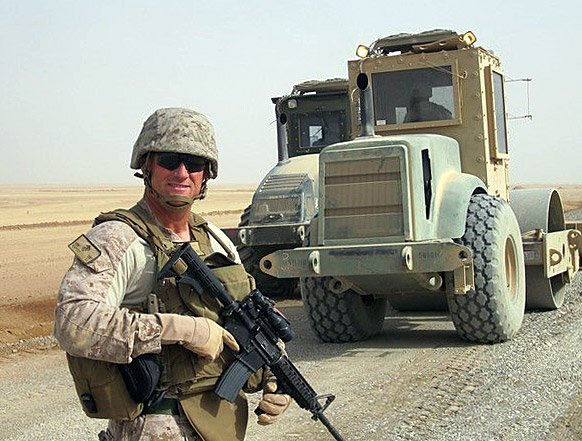 US Marine in uniform holding rifle with two military trucks in background