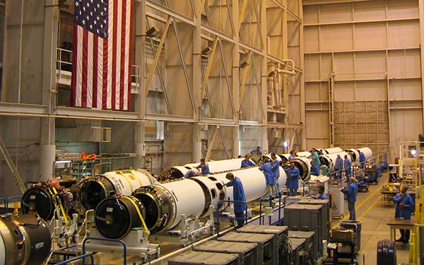 Workers in factory setting working on missile laying horizontal in the hanger. American flag hanging in the background