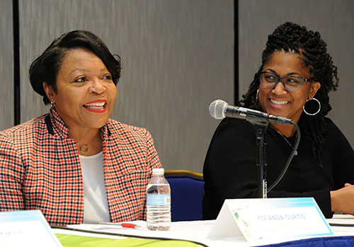 Two Black women speaking at a table in front of a microphone